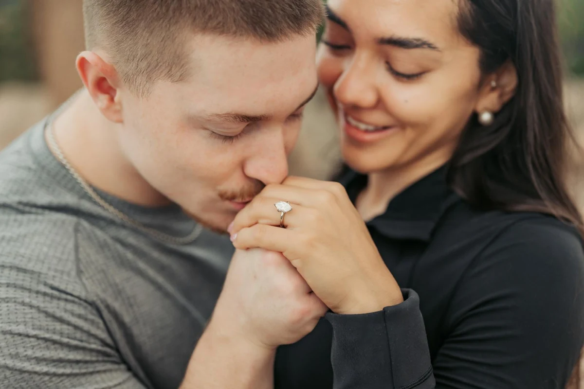An Ovoeu couple holding hands, with one partner kissing the other's hand beside an Ovoeu custom Dutch Marquee Solitaire ring.